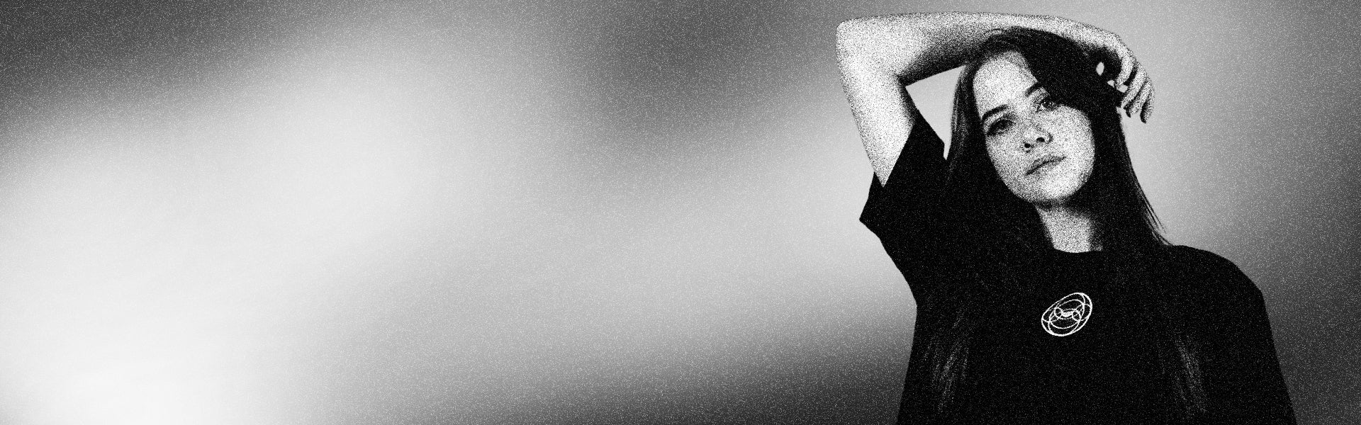 Black-and-white photograph capturing a woman with long hair in a dynamic, energetic pose, one arm raised, set against a gradient background that evokes a sense of movement and progress. Her dark shirt features an abstract circular design, symbolizing creativity and innovation. The image’s grainy, artistic texture adds a modern, forward-thinking edge.